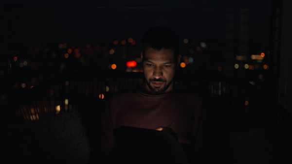 A young African AMerican man sitting on balcony with urban view and using tablet at night