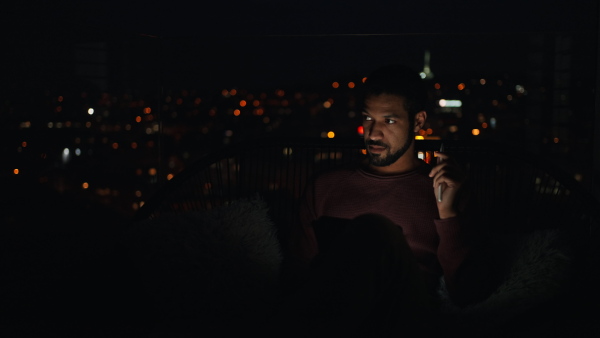 A young African AMerican man sitting on balcony with urban view and using tablet at night