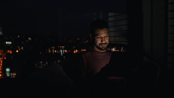 A young African AMerican man sitting on balcony with urban view and using tablet at night