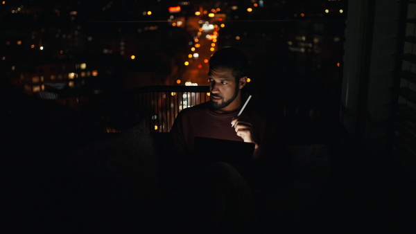 A young African AMerican man sitting on balcony with urban view and using tablet at night