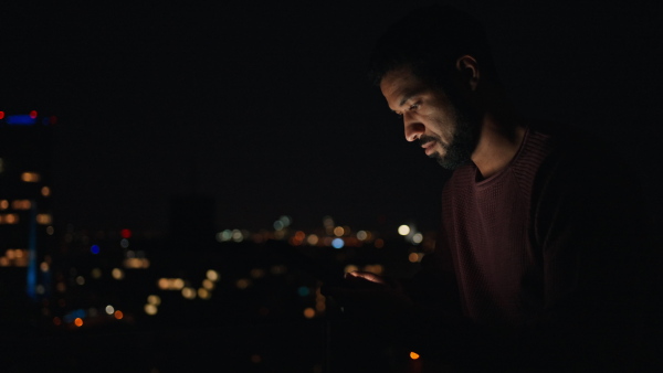 A young African AMerican man sitting on balcony with urban view and using tablet at night