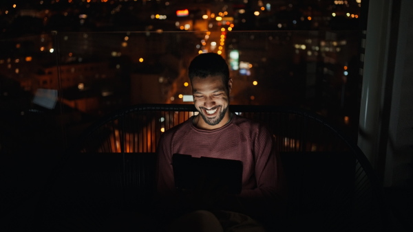 A young African AMerican man sitting on balcony with urban view and having video call at night