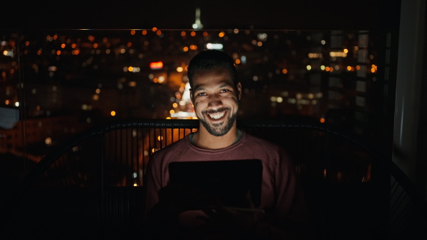 A young African AMerican man sitting on balcony with urban view and using tablet at night