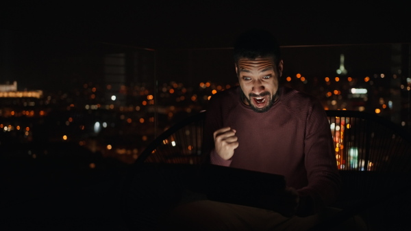 A young African AMerican man sitting on balcony with urban view and using tablet at night