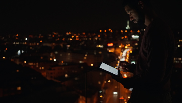 A young African AMerican man sitting on balcony with urban view and using tablet at night.