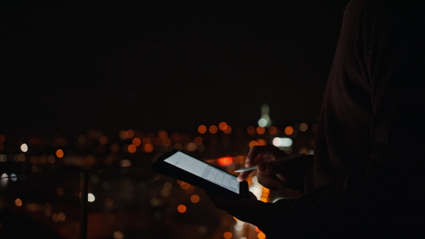 A young African AMerican man sitting on balcony with urban view and using tablet at night, close-up