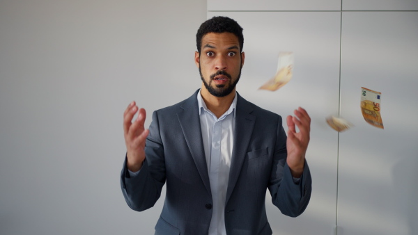 A serious young businessman man standing in office with money falling down around him, inflation concept.