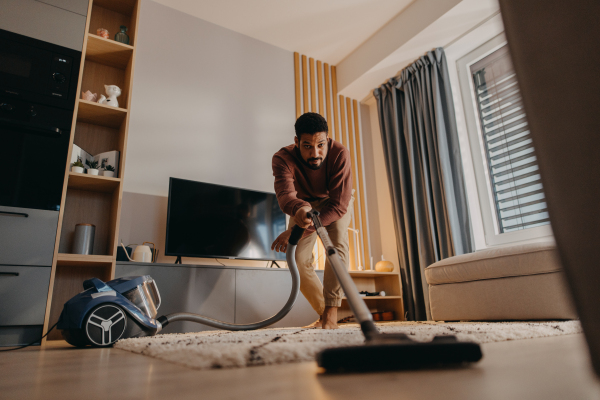 A young man hoovering carpet with vacuum cleaner in living room