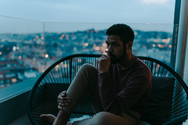 A portrait od depressed young African - American man sitting on balcony with urban view in evening.