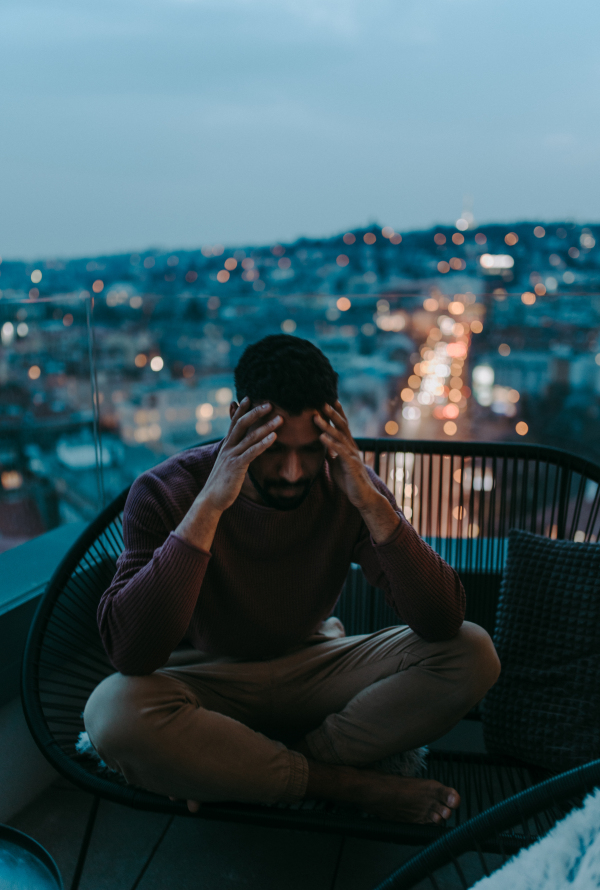 A portrait od depressed young African American man with head in hands sitting on balcony at night.