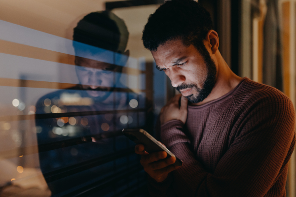 A young man with a smart phone looks worried, standing at balcony in evening at home.