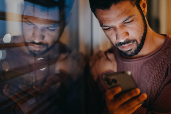 A young man with a smart phone looks sad, standing by window in evening at home.