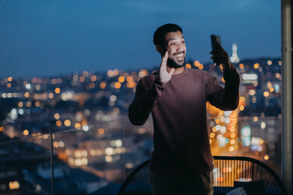 A cheerful young man taking selfie on balcony with urban view at night.