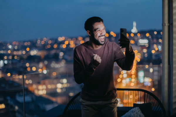 A cheerful young man taking selfie on balcony with urban view at night.