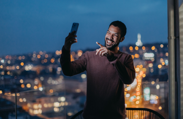 A cheerful young man taking selfie on balcony with urban view at night.