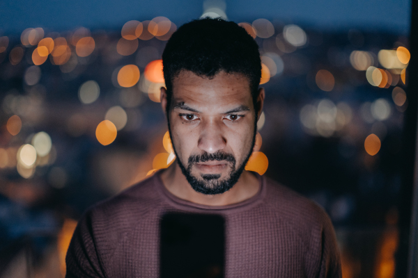 A young man with a smart phone looks worried, standing at balcony with city view in evening at home.