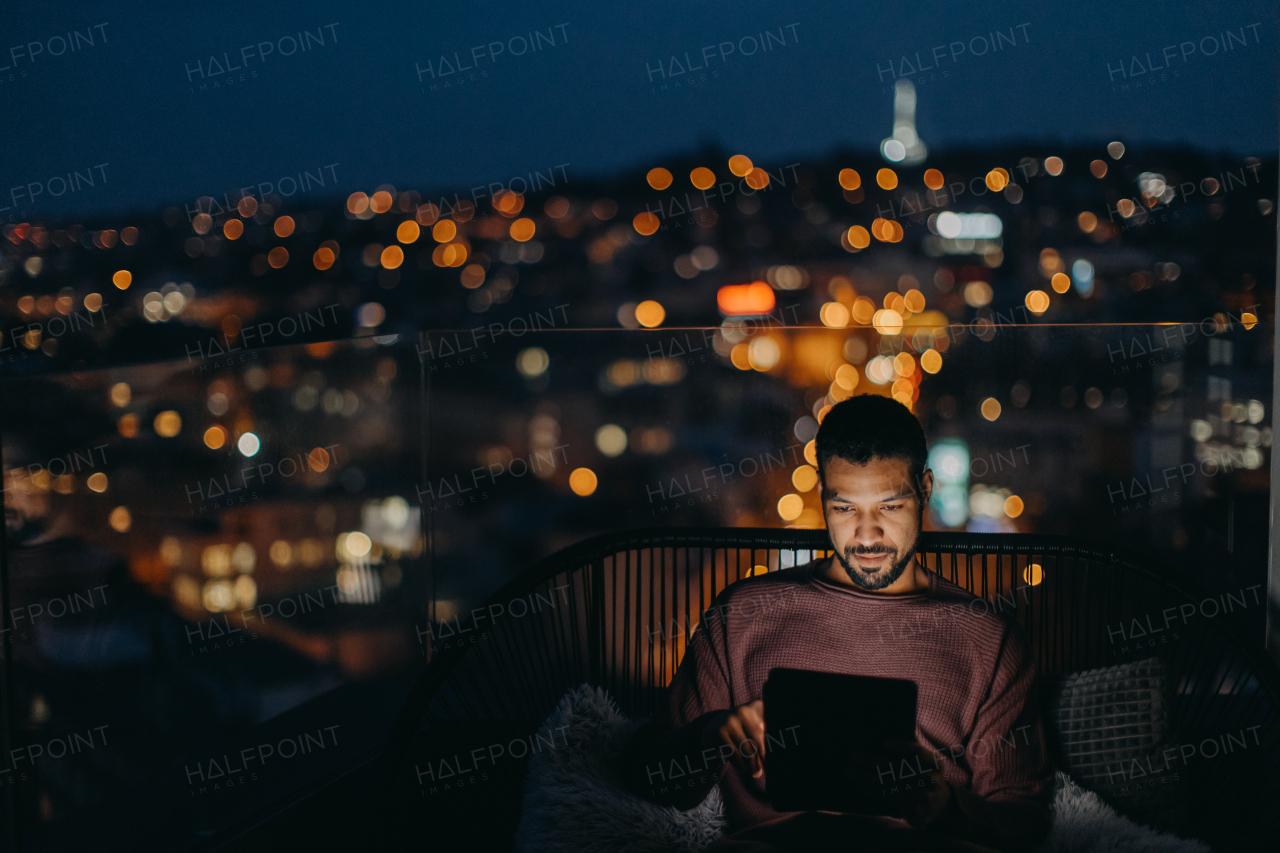 A young African AMerican man sitting on balcony with urban view and using tablet at night