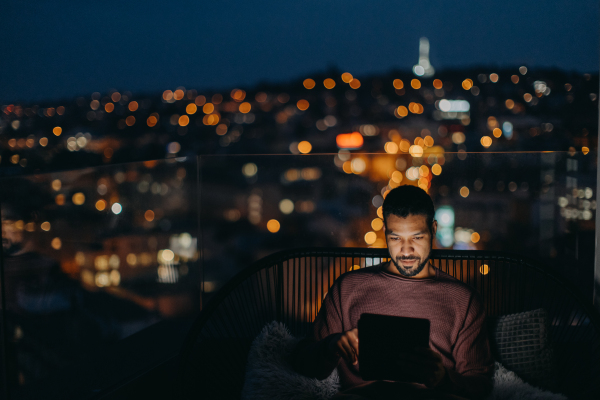 A young African AMerican man sitting on balcony with urban view and using tablet at night