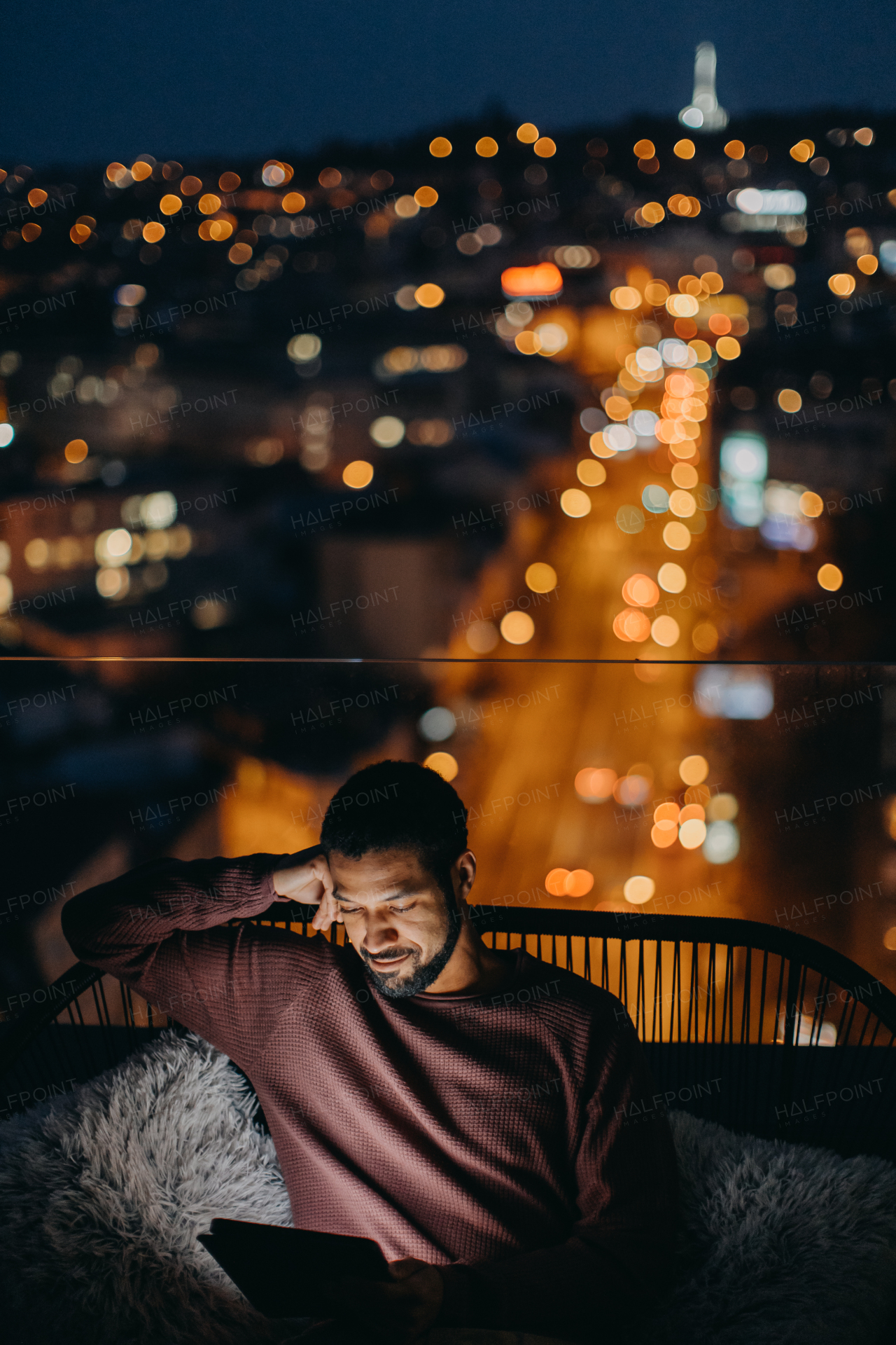 A young African AMerican man sitting on balcony with urban view and using tablet at night