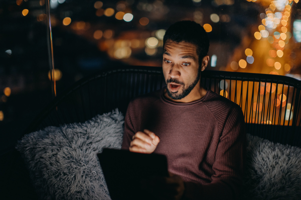 A young African AMerican man sitting on balcony with urban view and using tablet at night