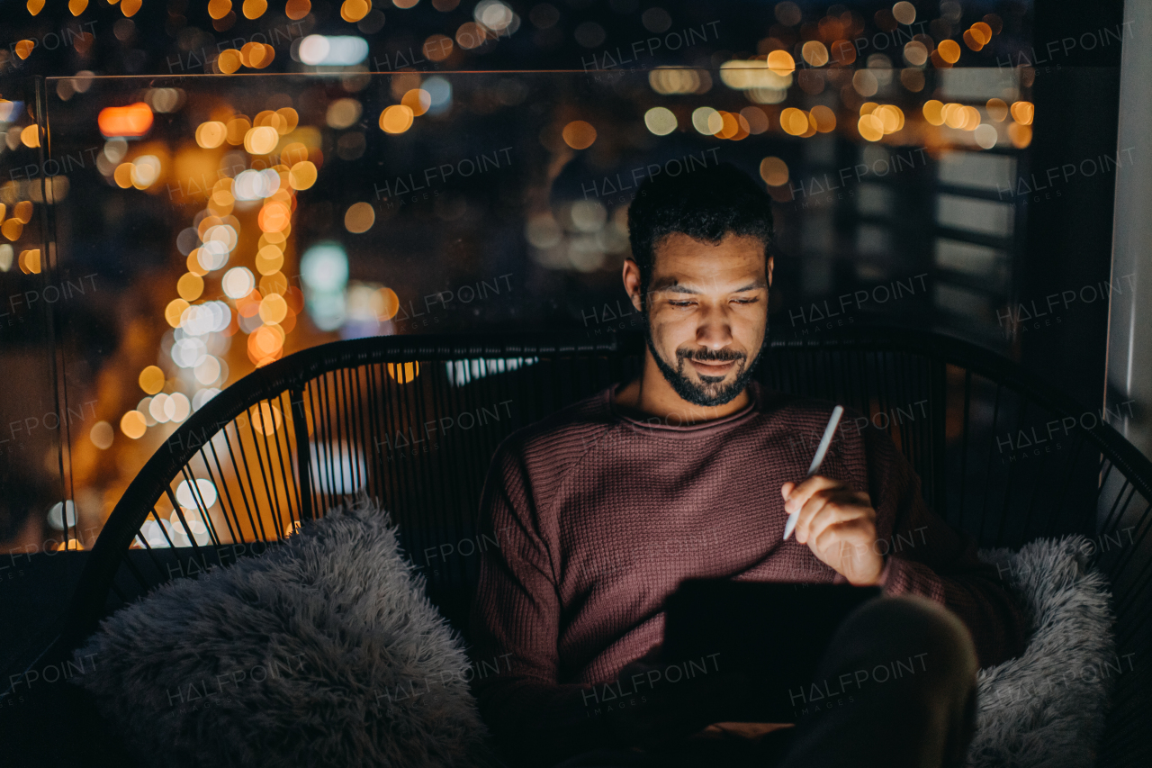 A young African AMerican man sitting on balcony with urban view and using tablet at night