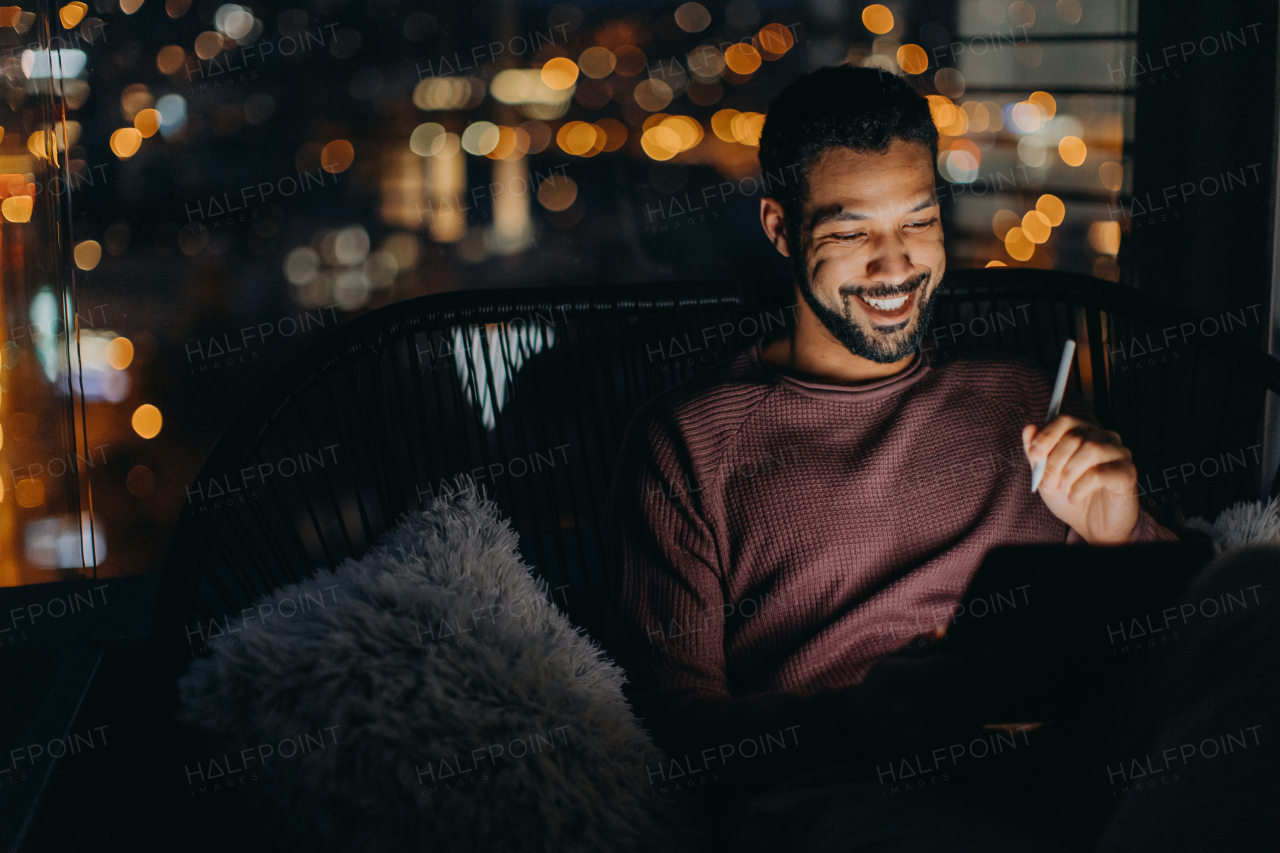 A young African AMerican man sitting on balcony with urban view and using tablet at night