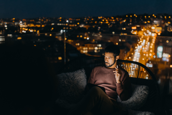 A young African AMerican man sitting on balcony with urban view and using tablet at night