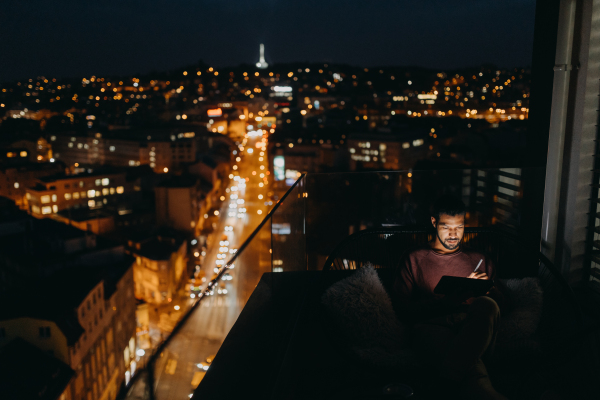 A young African AMerican man sitting on balcony with urban view and using tablet at night