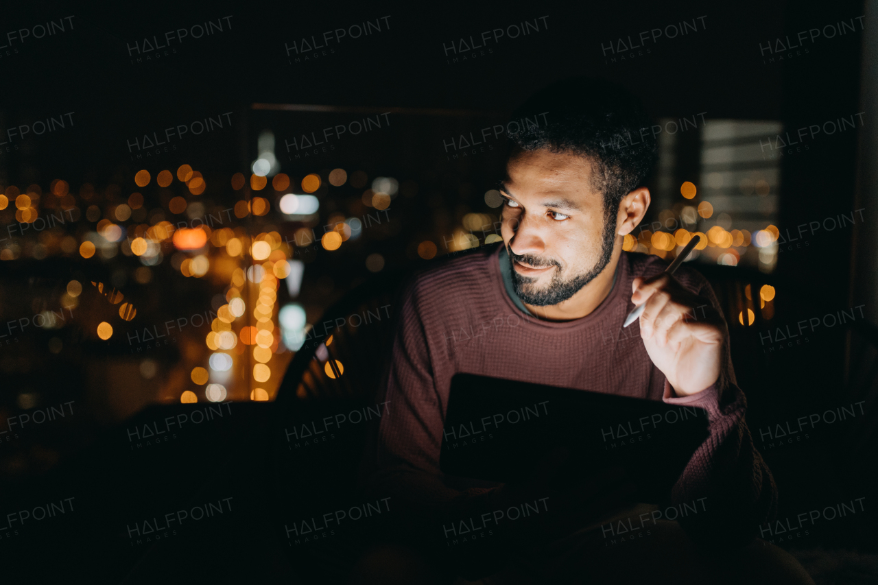 A young African AMerican man sitting on balcony with urban view and using tablet at night