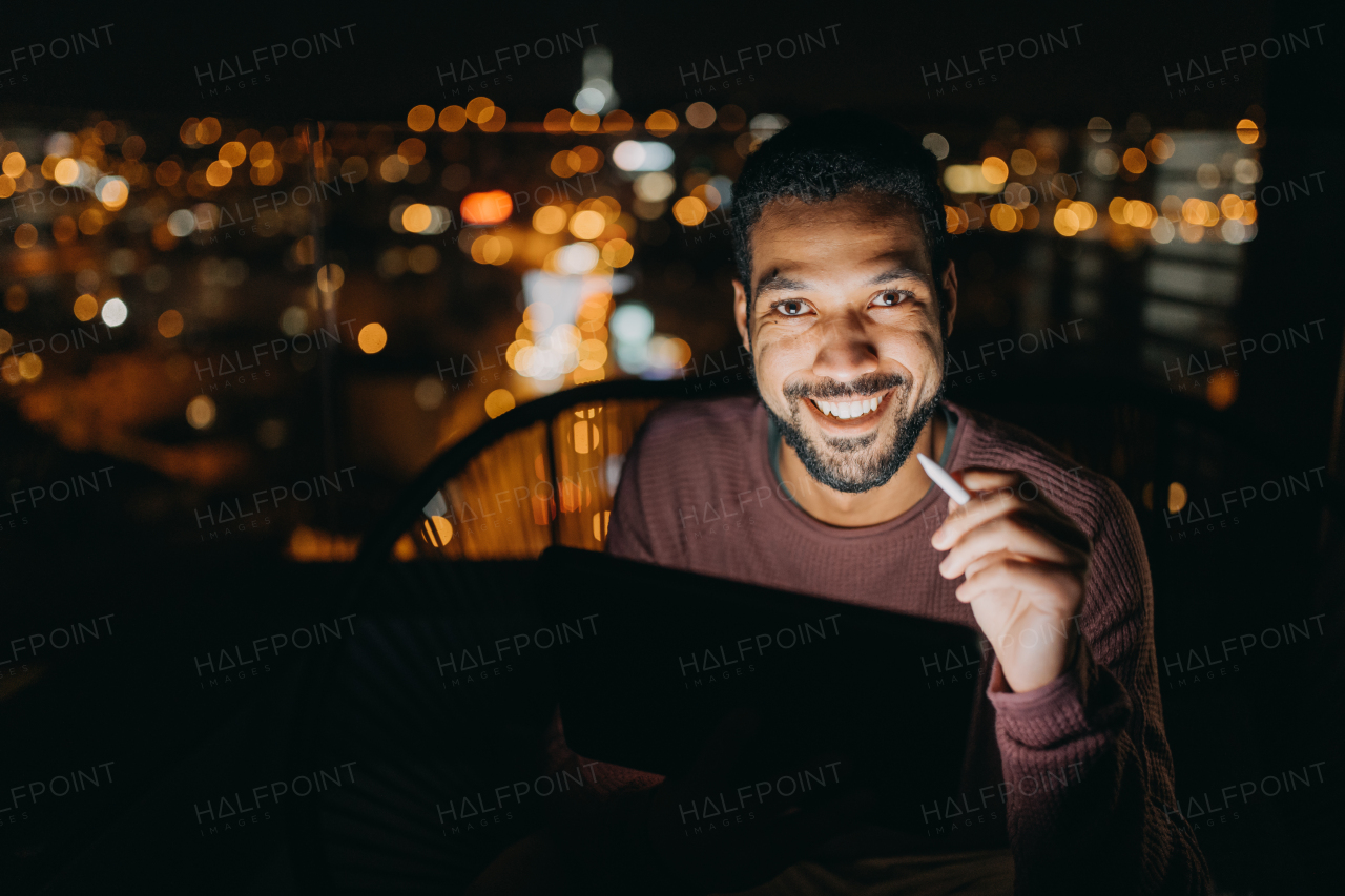 A young African AMerican man sitting on balcony with urban view and using tablet at night