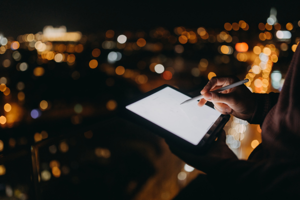 a Close-up of man standing on balcony with urban view and using tablet at night