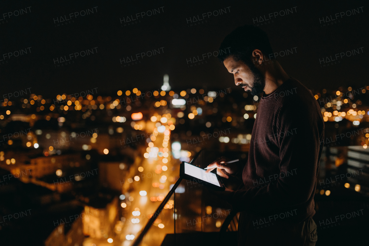 A young African American man standing on balcony with urban view and using tablet at night