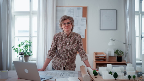 Happy senior woman architect with model of houses standing in office and looking at camera.