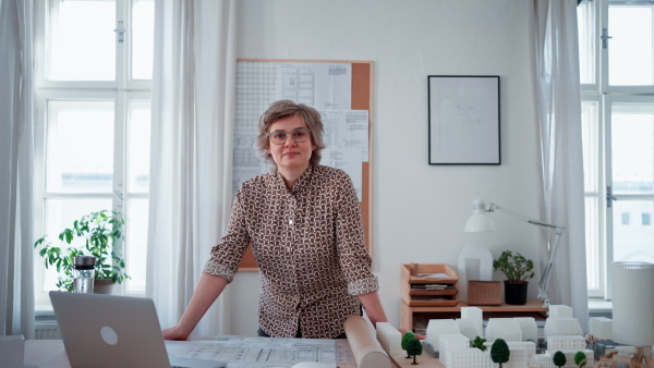 Happy senior woman architect with model of houses standing in office and looking at camera.