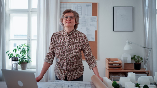 Happy senior woman architect with model of houses standing in office and looking at camera.