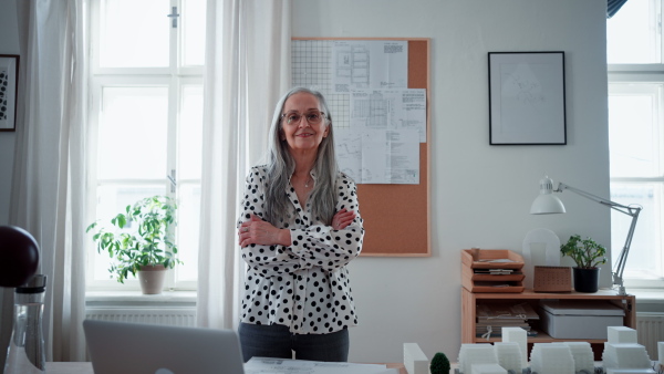 Happy senior woman architect with model of houses standing in office and looking at camera.