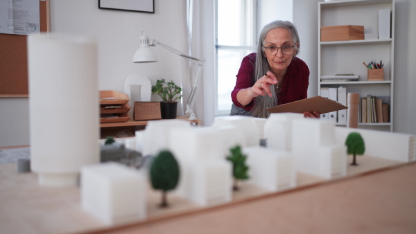 A senior woman eco architect with model of modern bulidings working in office.