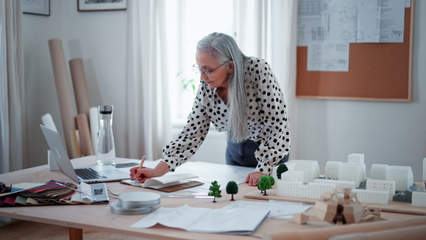 A senior woman eco architect with model of modern bulidings working in office.