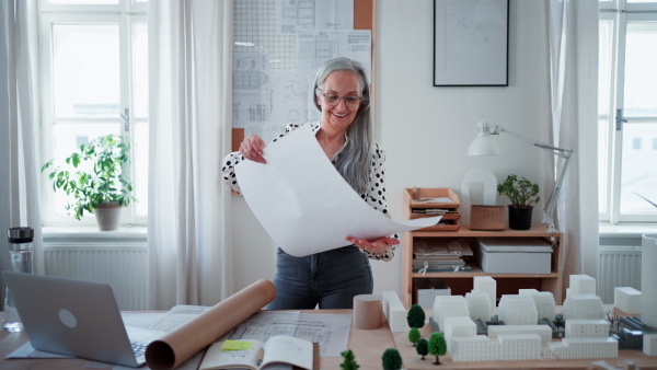 A senior woman architect with model of houses looking at blueprints in office.