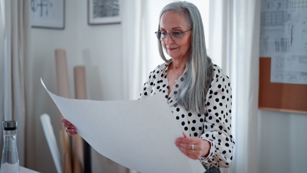 A senior woman architect with model of houses looking at blueprints in office.