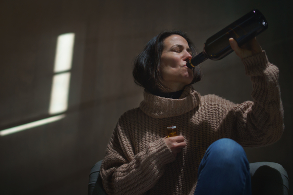 A depressed drunk woman sitting on the floor in the dark taking pills and drinking wine from a bottle