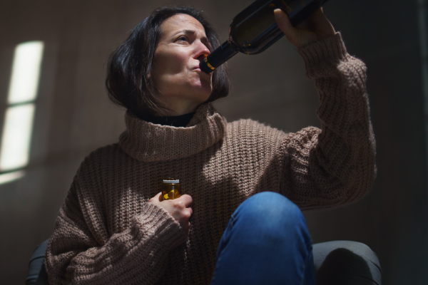 A depressed drunk woman sitting on the floor in the dark taking pills and drinking wine from a bottle