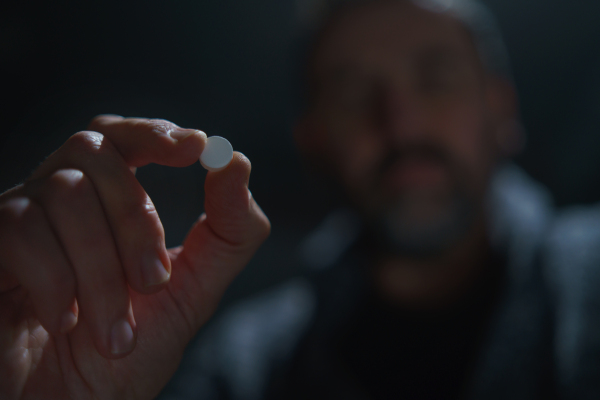 A blurred man holding and show drug pill on dark background, mental health concept.