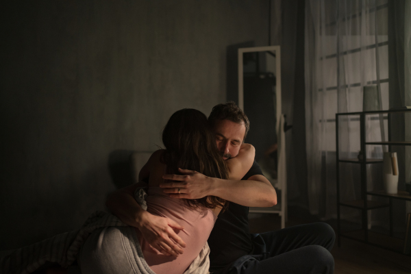 A mid-adult couple sitting on bed at night and hugging.