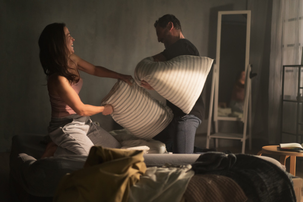 A happy couple beating with pillows on the bed in a bedroom at home.