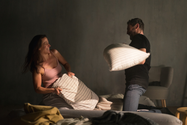 A happy couple beating with pillows on the bed in a bedroom at home.