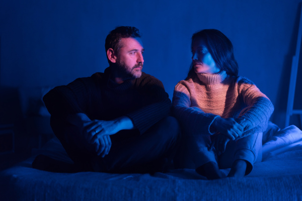 A sad mid-adult couple sitting on bed at night, looking at camera, suffering from depression after covid.