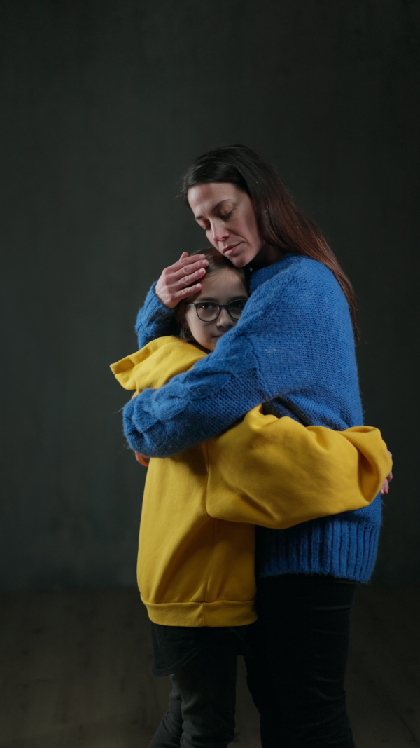 A sad refugee mother hugging her daughter, both wearing Ukrainian national colors on black background.