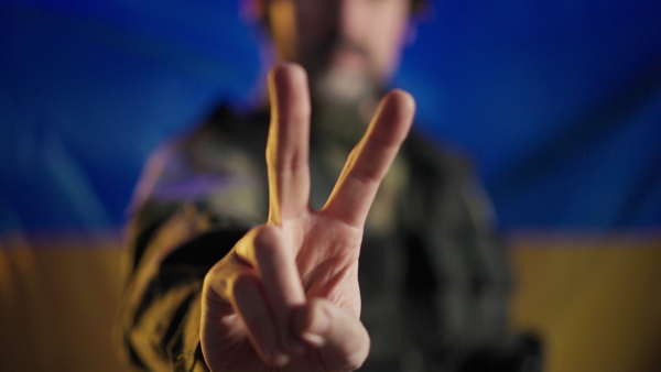 A soldier in military uniform showing peace sign with Ukraine flag in background.