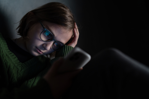 A sad little girl, alone in darkness, sitting and using smartphone.